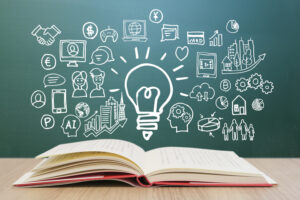 Open book on a table with a chalkboard full of lightbulb and business icons, representing ideas and learning.