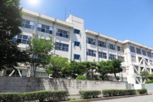 White multi-story school building with large windows, set behind a concrete wall and green trees under a clear blue sky