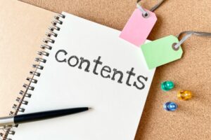 Notebook page titled 'contents' with a black pen, pink and green tag labels, and colorful push pins on a corkboard backdrop.