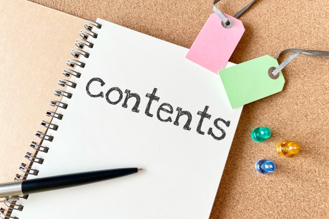 Notebook page titled 'contents' with a black pen, pink and green tag labels, and colorful push pins on a corkboard backdrop.