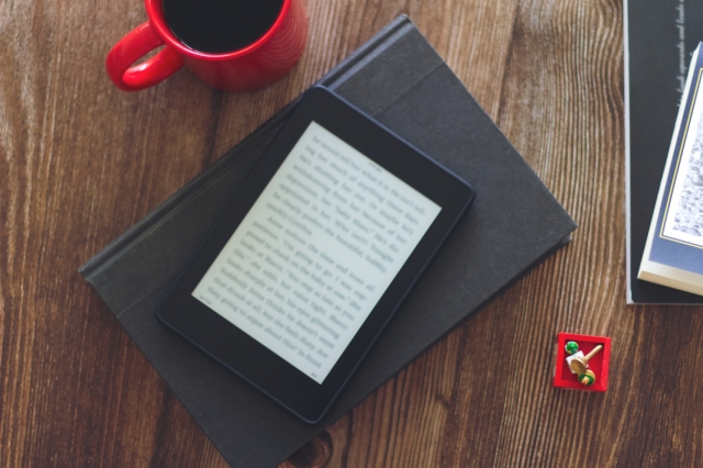 E-reader resting on a closed black notebook on a wooden desk, with a red mug nearby and a small red toy block nearby.