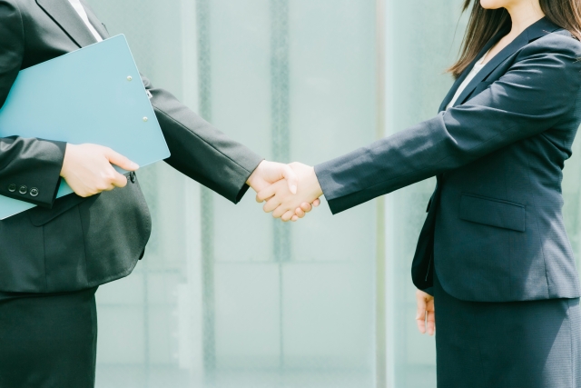 Two professionals in business suits shake hands, one holding a light blue folder, outside an office.
