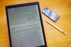 Tablet showing a page of dense text with red handwritten marks on a wooden desk, beside a smartphone and a white stylus/pen.