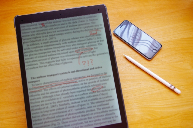 Tablet showing a page of dense text with red handwritten marks on a wooden desk, beside a smartphone and a white stylus/pen.