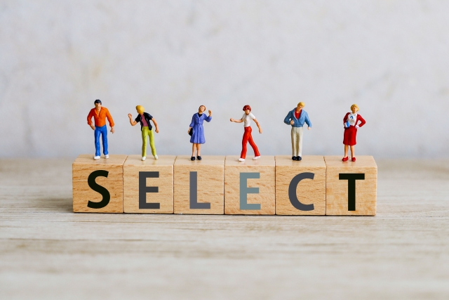 Miniature figures stand on wooden blocks spelling SELECT, illustrating choosing or selection among people.