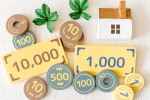 Pile of colorful play money notes and round coins with a small white house figurine, suggesting savings or real estate investment.