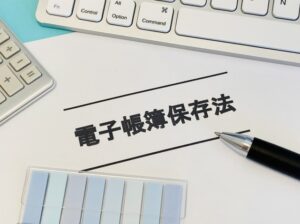 Close-up of a document titled '電子帳簿保存法' (Electronic bookkeeping retention law) with a pen nearby, a keyboard and calculator in view, and color swatches nearby.