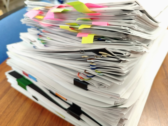 Large stack of documents covered in colorful sticky notes on a wooden desk, suggesting heavy paperwork or filing.