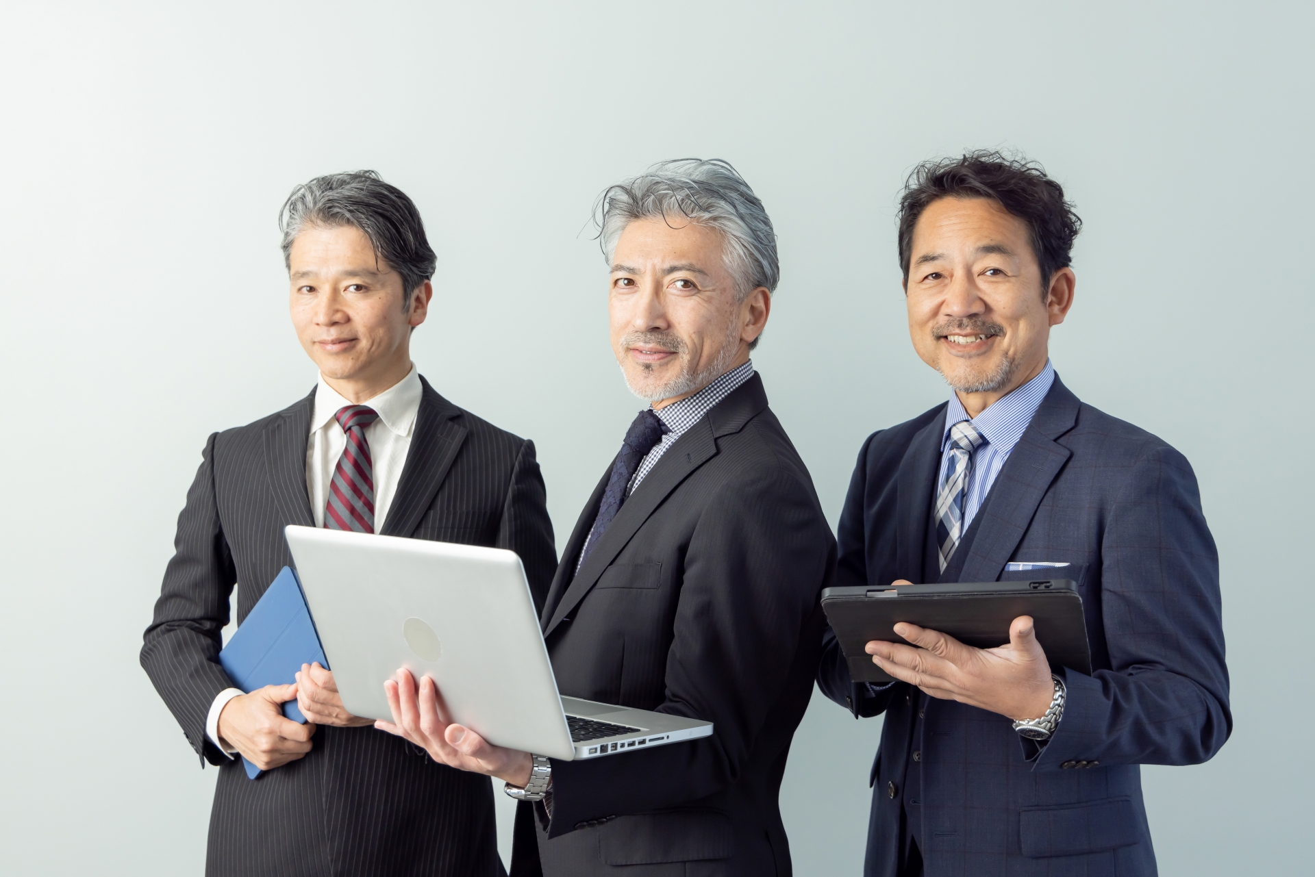Three businessmen in suits pose with devices: a laptop and blue folder in the left, a laptop in the middle, and a tablet in the right.