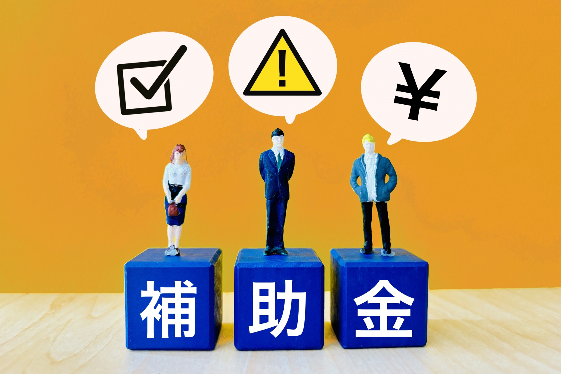 Three figurines stand on blue blocks that spell 補助金, with speech bubbles showing a checkmark, a warning triangle, and a yen symbol against an orange background.