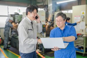Two factory workers discuss over a laptop, one on the phone, amid machinery and sparks in a workshop.