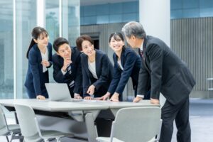 Five professionals in business attire lean over a conference table, looking at a laptop during a collaborative meeting outdoorsy office setting.