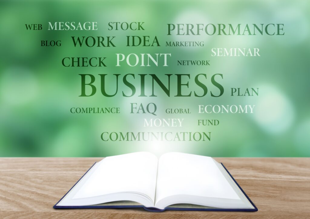 Open book resting on a wooden desk with a green blurred background of business words floating above it.