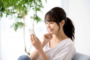 Woman sitting on a sofa, smiling at her smartphone held in her right hand, in a bright room with a plant in the background.