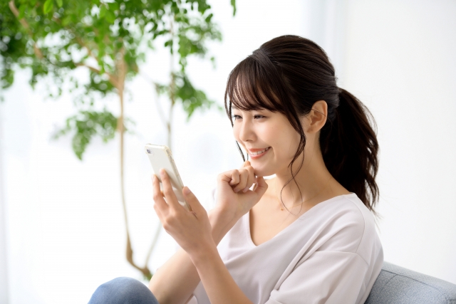 Woman sitting on a sofa, smiling at her smartphone held in her right hand, in a bright room with a plant in the background.
