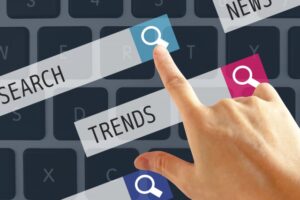 Hand taps a blue sticky note with a magnifying-glass icon on a keyboard, signaling online search and trends.